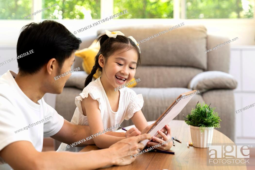 Stock Photo: Asian girl reading tale story book with her father in living room while city lockdown from coronavirus covid-19 pandemic.