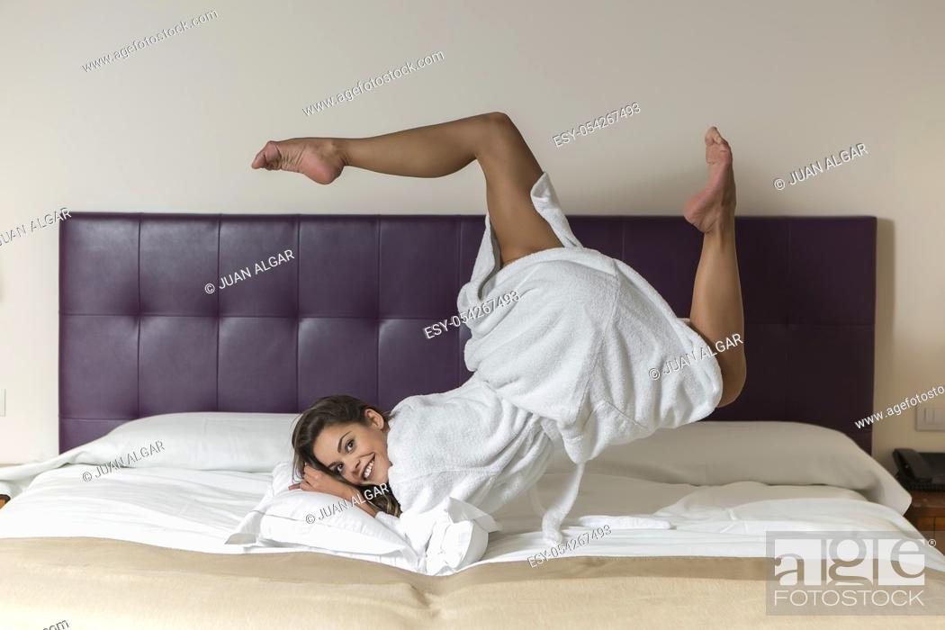 Stock Photo: Attractive young female in white bathrobe rolling in bed in hotel room.