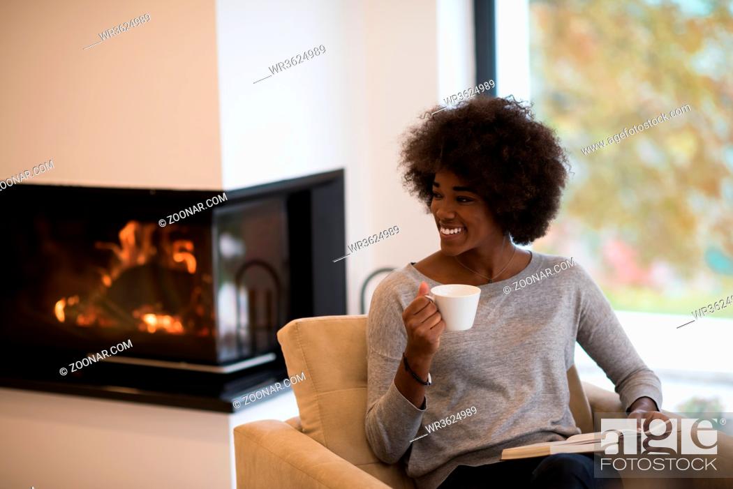 Stock Photo: african american woman drinking cup of coffee reading book at fireplace. Young black girl with hot beverage relaxing heating warming up.