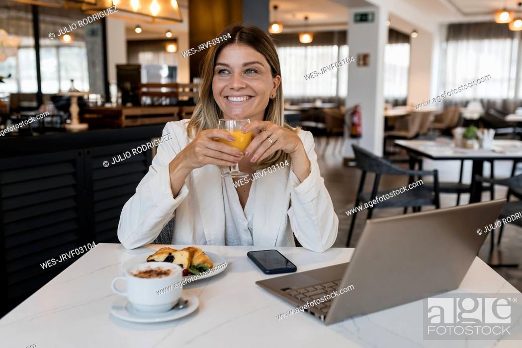Imagen: Smiling businesswoman holding juice glass sitting with laptop at restaurant.