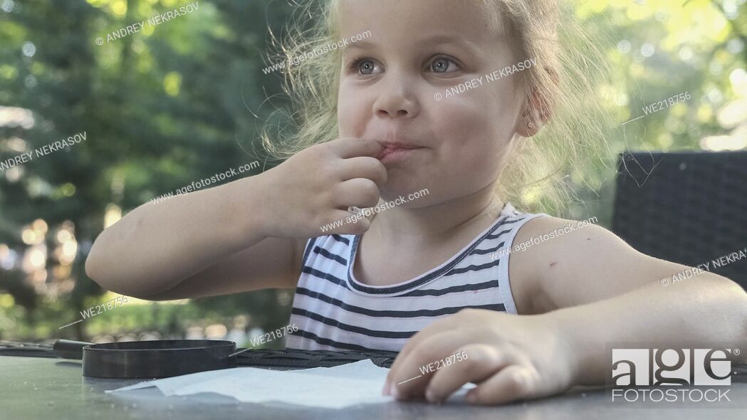Stock Photo: Little girl tastes salt. Close-up portrait of blonde girl takes salt from napkin with her finger and tastes it while sitting in street cafe on the park.