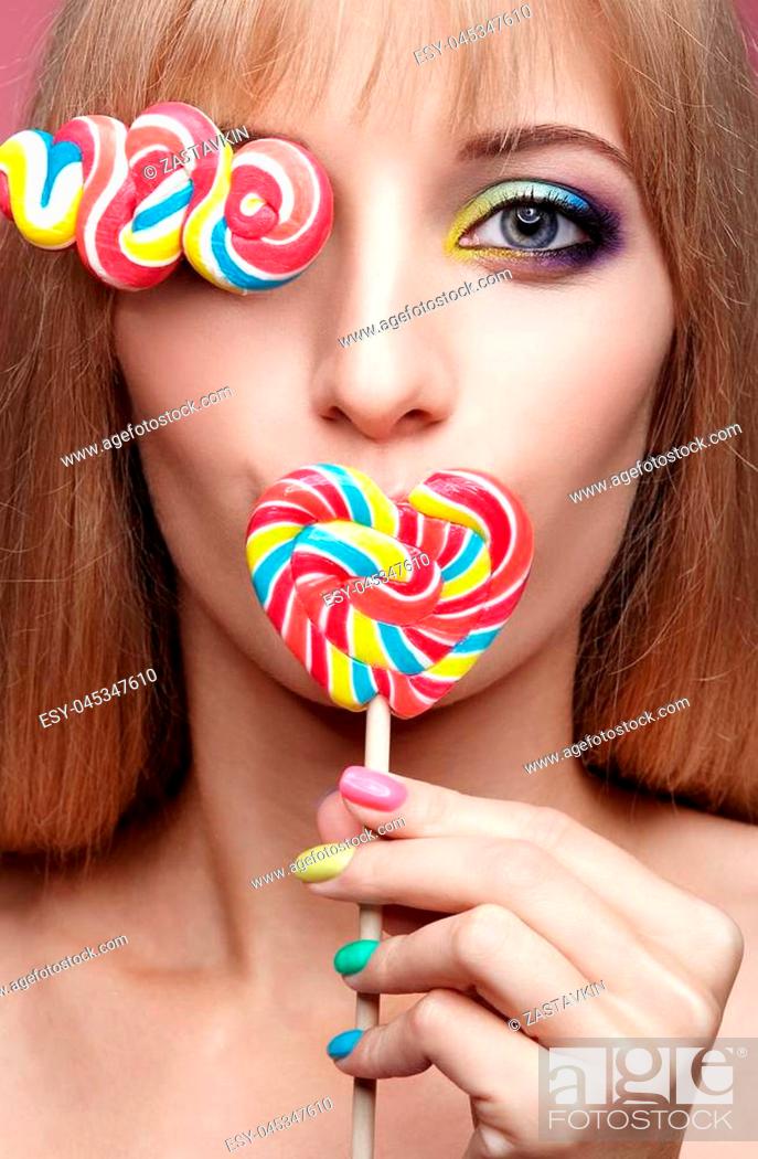 Stock Photo: Beauty portrait of young blonde woman on pink background. Female with candy lollipop on stick in hands. Girl has finger nails with bright yellow, green.
