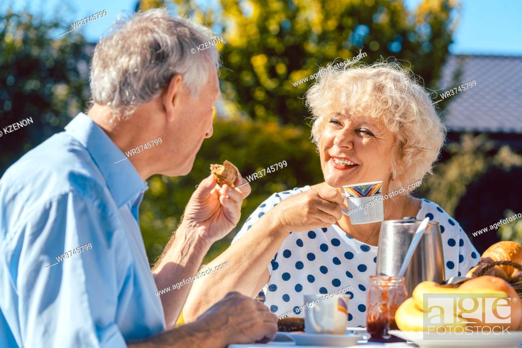 Stock Photo: Happy elderly woman and man eating breakfast sitting in their garden outdoors in summer, eating bread rolls and drinking coffee.