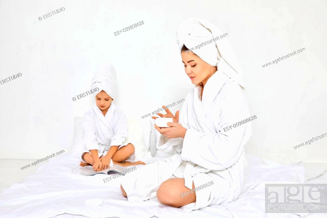 Imagen: happy caucasian mother in white bathrobes and towels on head apply cream on hands and her daughter reading magazine on background.