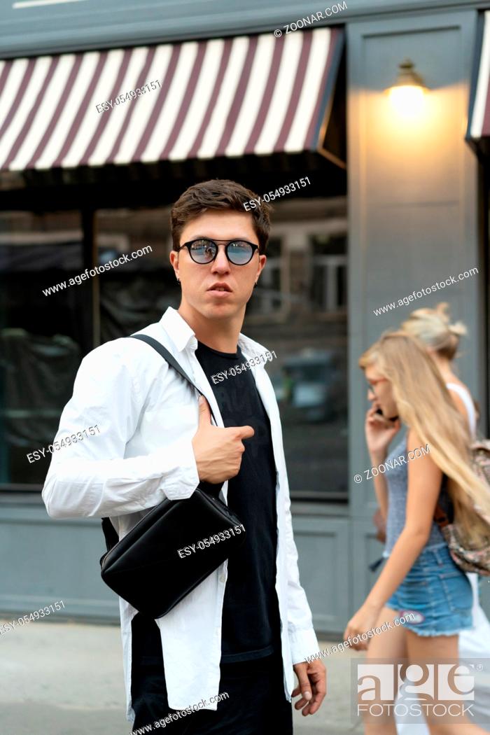 Stock Photo: Young guy in round sunglasses posing on camera. A small bag hangs over his shoulder.