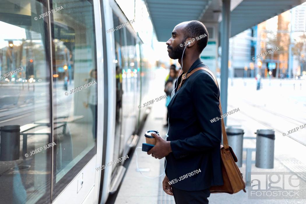 Stock Photo: Young businessman with coffee to go standing at tram stop listening music with earphones and smartphone.