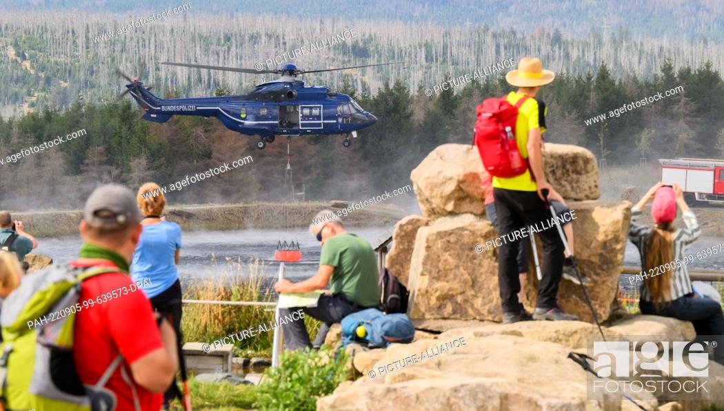Stock Photo: 05 September 2022, Lower Saxony, Braunlage: A helicopter of the federal police takes water from a lake on the Wurmberg, while numerous hikers and onlookers.