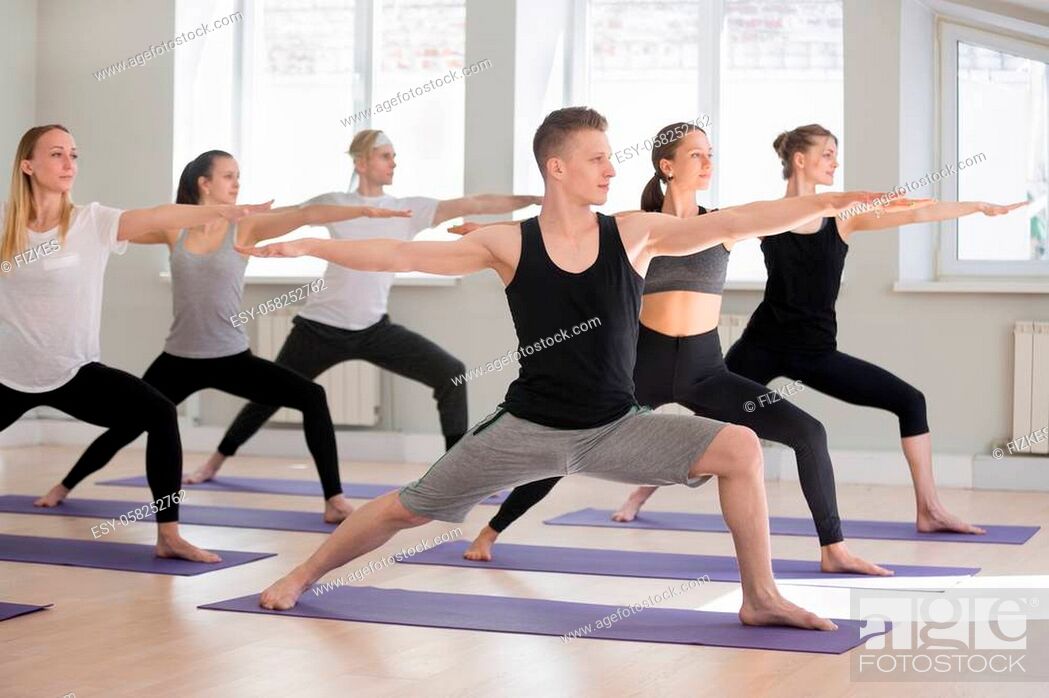 Stock Photo: Group of young attractive sporty people practicing yoga lesson, doing Warrior Two exercise, Virabhadrasana II pose, working out indoor full length.