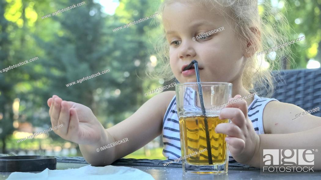 Stock Photo: Cute little girl drinks juice through straw. Close-up portrait of blonde girl drinks juice from glass through cocktail straw sitting in street cafe on the park.