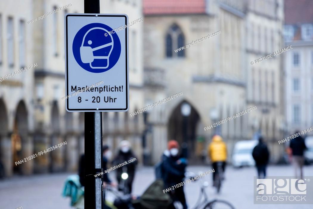 Stock Photo: 01 March 2021, North Rhine-Westphalia, Münster: People walk across the Prinzipalmarkt. A sign with the inscription ""Maskenpflicht! 8-20 o'clock"" indicates.