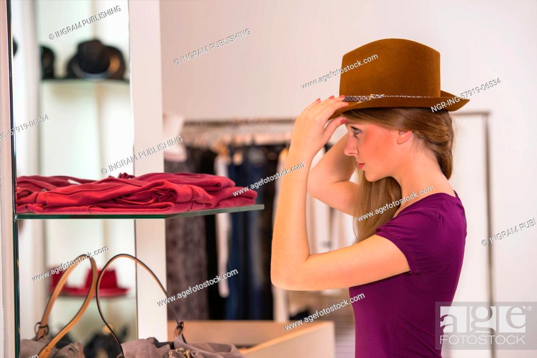 Stock Photo: Beautiful woman shopping in clothing store trying on brown hat in front of mirror.