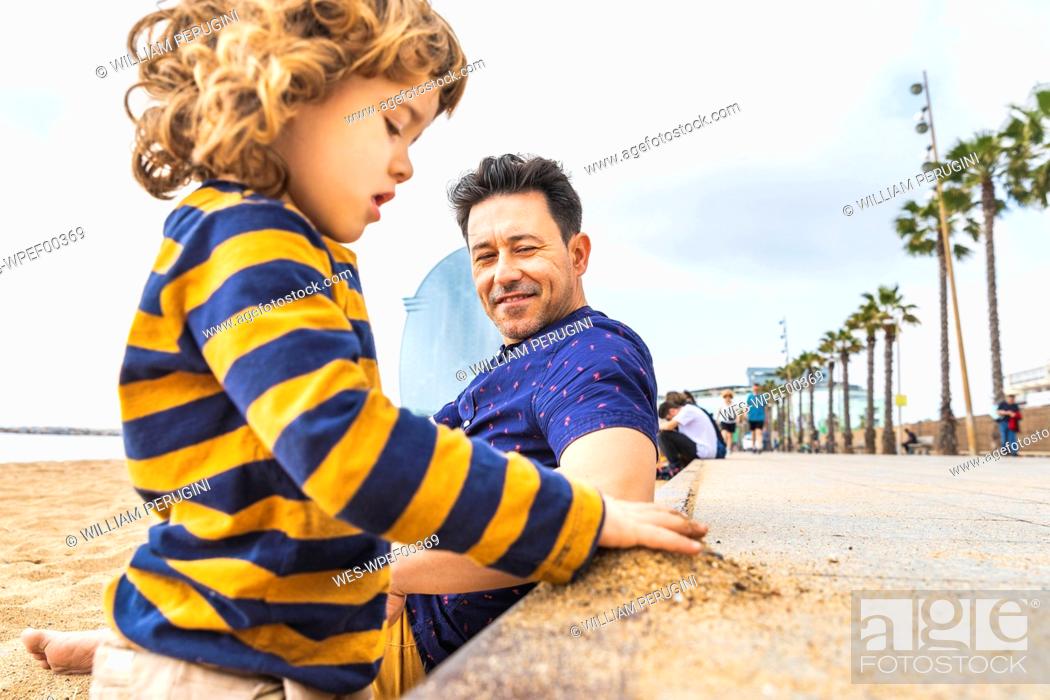 Stock Photo: Spain, Barcelona, young boy playing with sand, his father sitting next to him and smiling.
