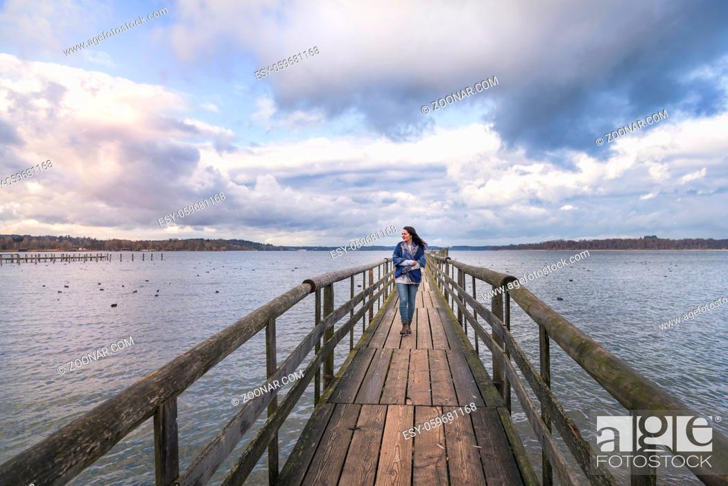 Stock Photo: Beautiful young woman smiling and walking alone on a long wooden pontoon, over the lake Chiemsee, enjoying the fresh air and the sunset, in Germany.
