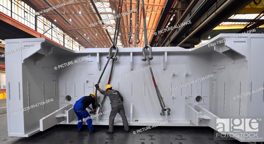 Stock Photo: 20 February 2019, Thuringia, Erfurt: Two employees fasten the component of a large press to the crane in a production hall of Schuler Pressen GmbH.