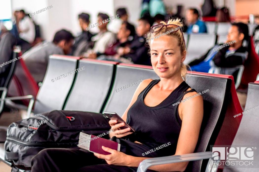 Stock Photo: Casual tanned blond female traveler holding cell phone, passport and boarding pass while waiting to board a plane at the departure gates at the asian airport.