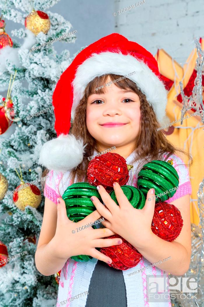 Stock Photo: Portrait of a laughing preschool girl in Santa hat holding Christmas decoration in hands.