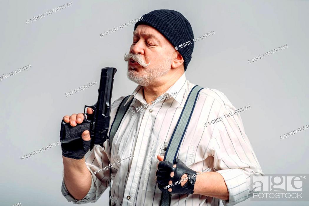 Stock Photo: Elderly man with gun isolated on grey background, gangster. Mature senior in hat holds weapon, robber in old age.