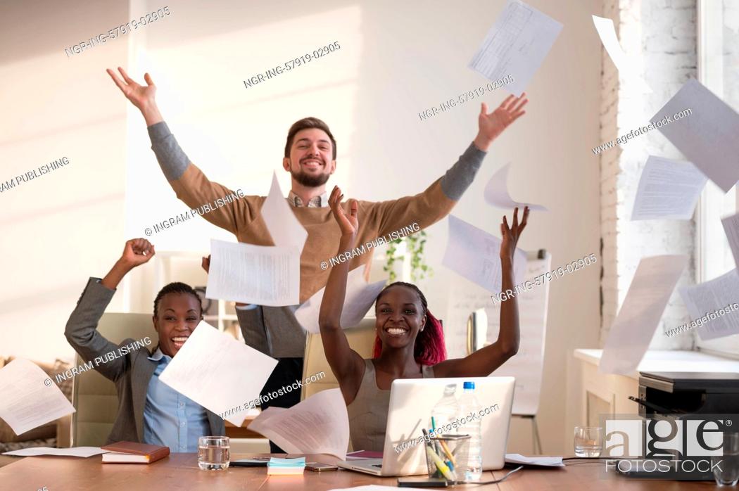 Stock Photo: Business people excited happy smile, throwing up papers, documents fly in air, businesspeople sitting at office desk hold hands arms up, success team concept.