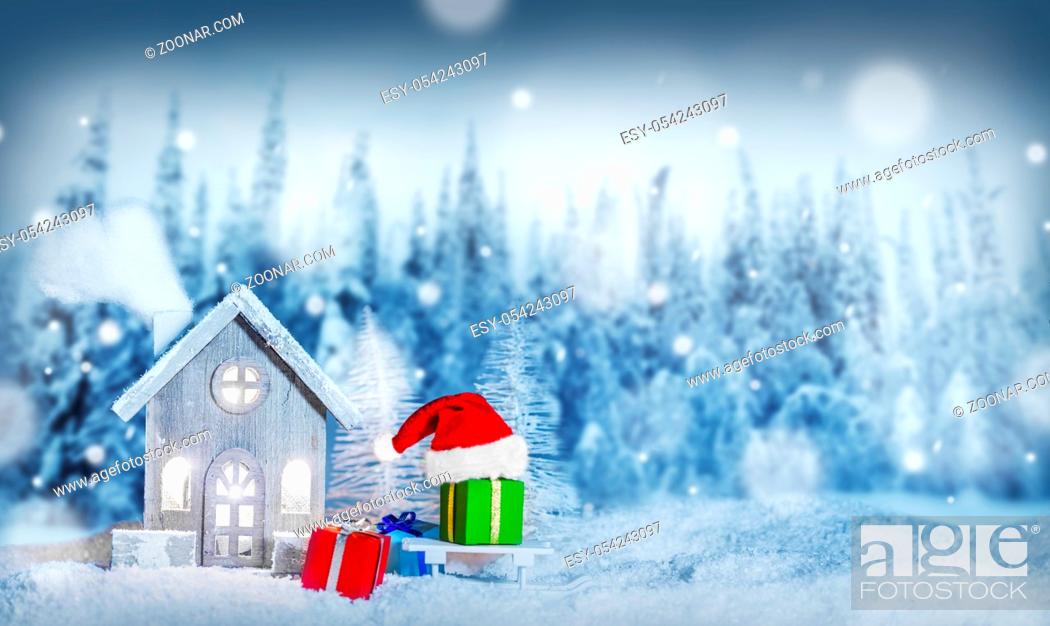 Stock Photo: Christmas card with cozy small decorative house , gifts and Santa Claus cap on sleigh on snow over fairy tale winter fir forest background and falling snow.
