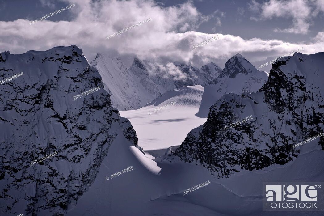 Stock Photo: Snow covered jagged mountain peaks with a purple-grey hue surrounding the sunlit glaciers under a cloud bank on the Juneau Ice Field in the Tongass National.