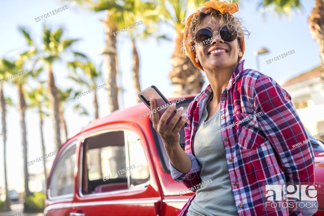 Photo de stock: Cheerful trendy young adult woman use app mobile phone to plan and set next travel destination traveling with a red classic car in background.