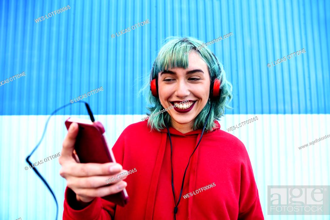 Stock Photo: Portrait of laughing young woman with blue dyed hair with headphones taking selfie with smartphone.