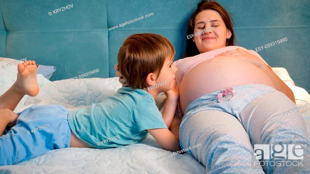 Stock Photo: Portrait of little toddler boy in pajamas lying with pregnant mother in bed and talking to unborn baby in big belly. Loving and caring children.