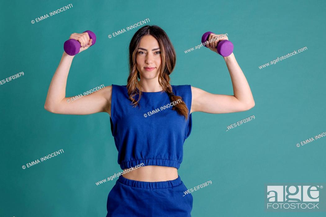 Stock Photo: Smiling young woman holding dumbbells flexing muscles against blue background.