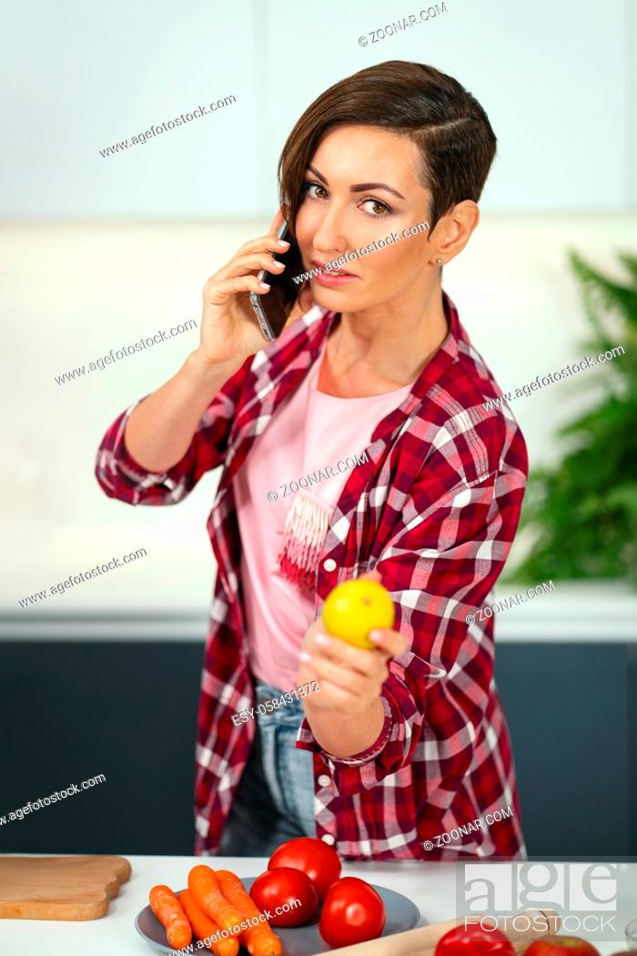 Stock Photo: Talking on the phone showing a lemon young pretty woman with a bob hairstyle prepares food in the kitchen. Healthy food at home.