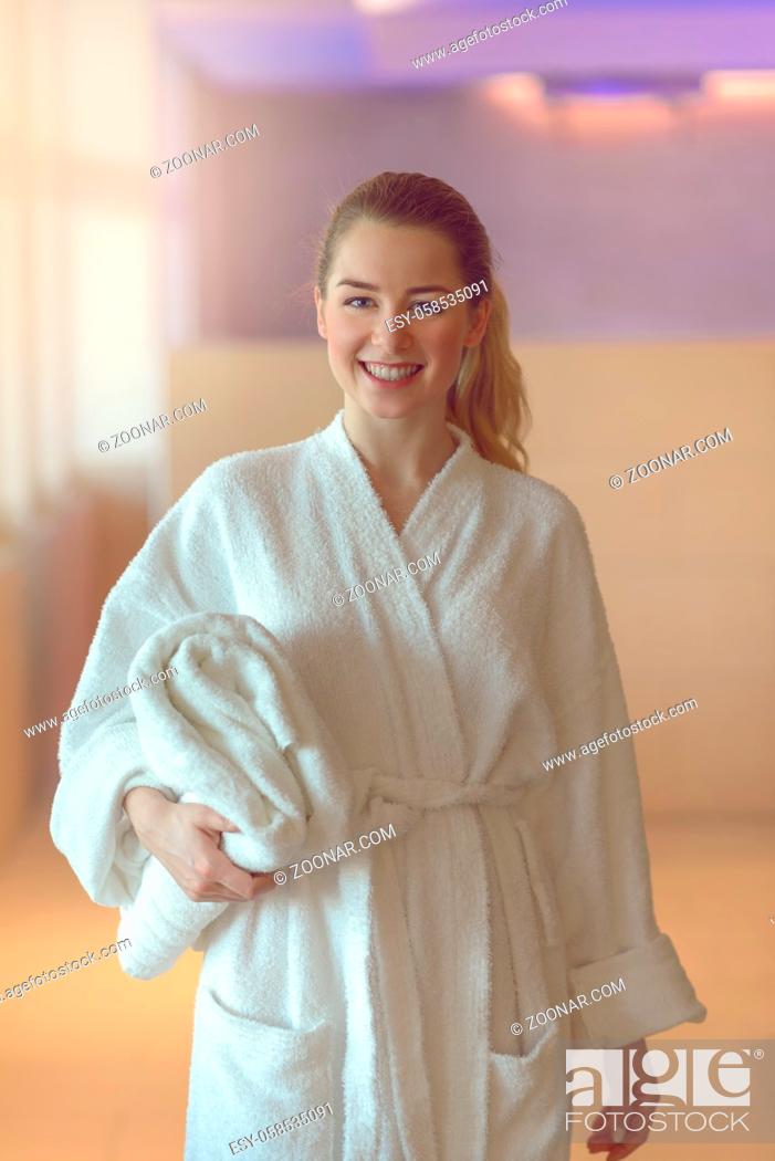 Imagen: Pretty young woman ready to take a bath or shower standing in a white towelling bathrobe holding a folded towel under her arm as she smiles at the camera.