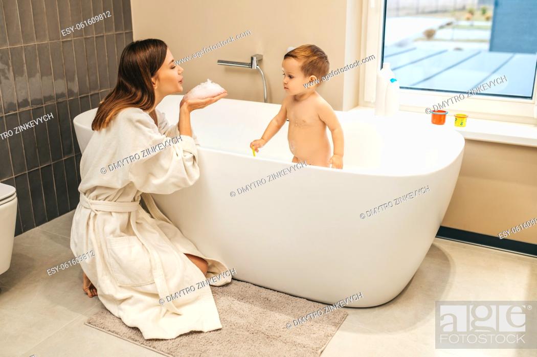 Stock Photo: Female in the bathrobe seated on her haunches blowing foam from her palms on the baby in the bath.