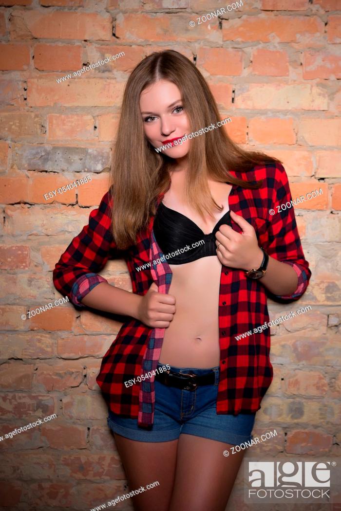 Stock Photo: Young smiling blond woman posing in checkered shirt and jeans shorts near the wall.