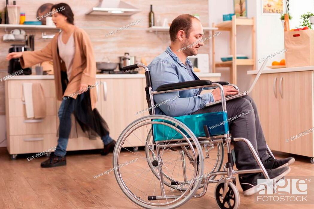 Stock Photo: Invalid man in wheelchair working remotely on laptop in kitchen while is standing in front of refrigerator. Disabled paralyzed handicapped man with walking.