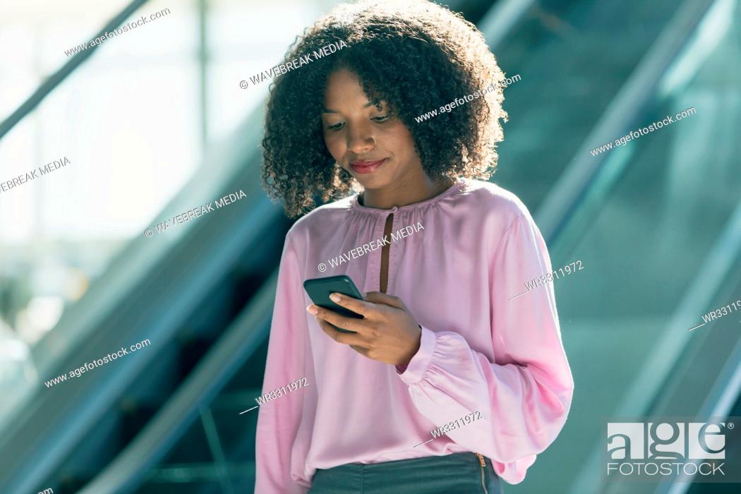 Stock Photo: African american businesswoman looking at mobile phone while using escalators in modern office.