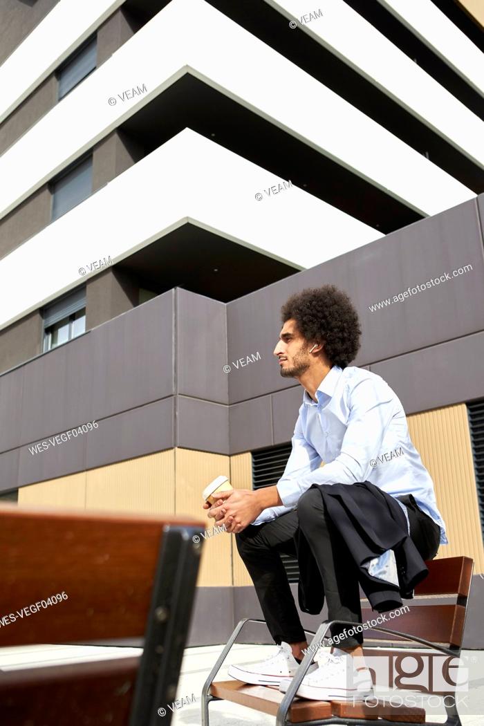 Stock Photo: Afro man with coffee cup looking away while sitting on chair in city.