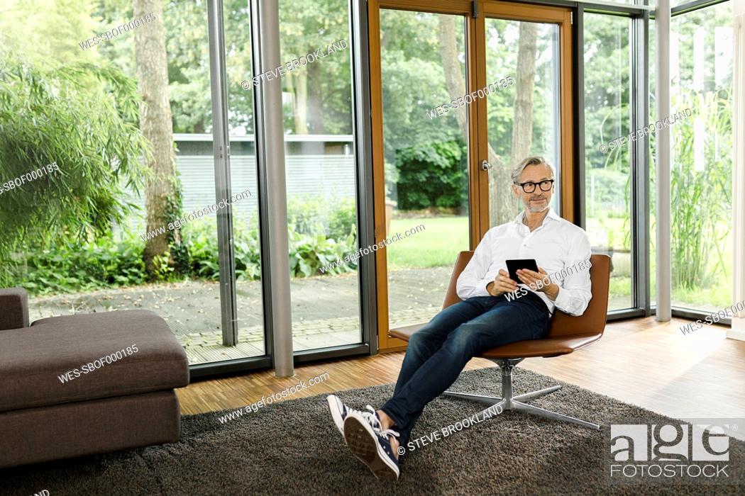 Stock Photo: Man sitting with tablet on chair in his living room.