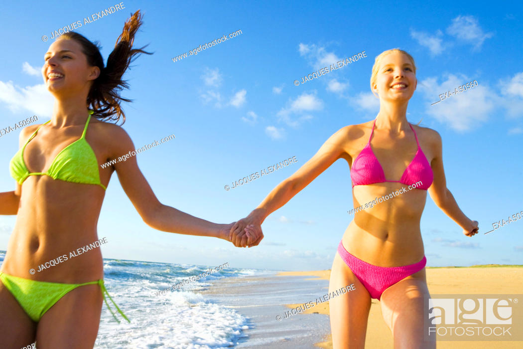Stock Photo: Two young women dressed in bikinis jogging along the coastal line and holding their hands.