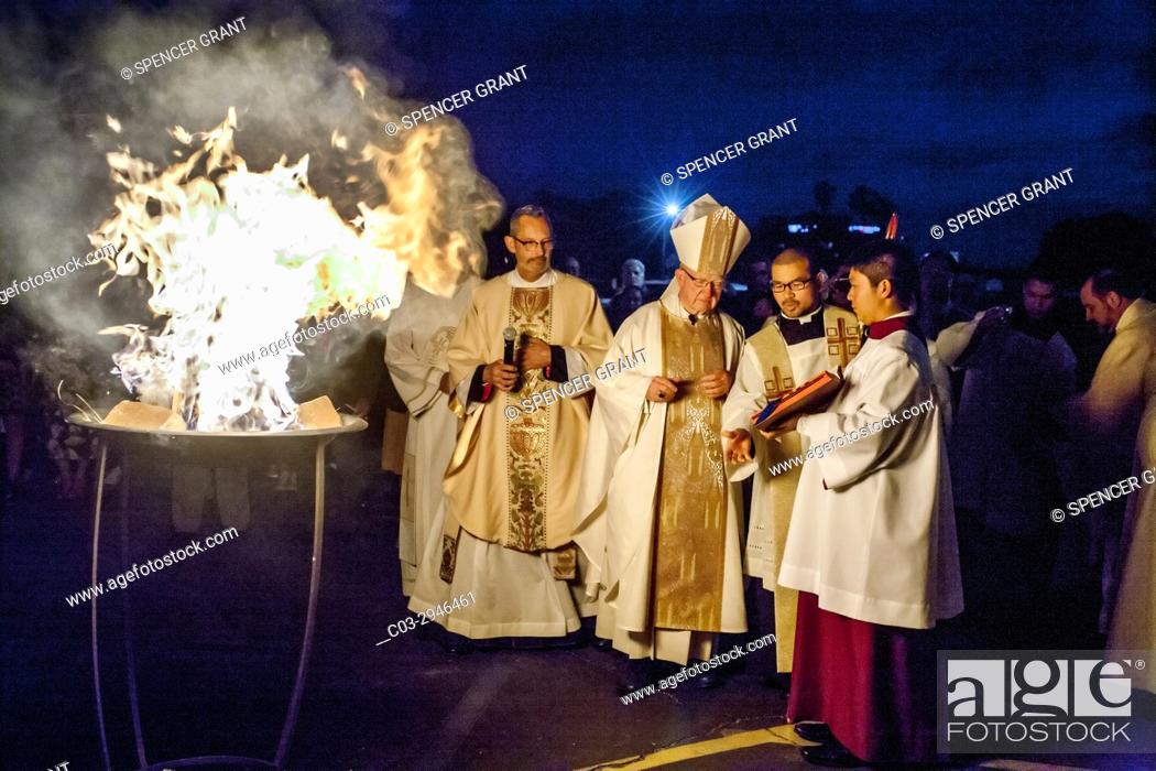 Stock Photo: By the light of the Easter fire, a Catholic bishop reads from the sacramentary book held by an Asian American pontifical server before conducting a christening.