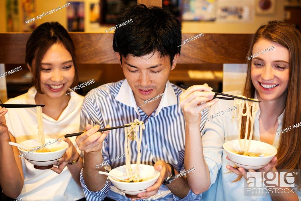 Photo de stock: Three smiling people sitting sidy by side at a table in a restaurant, eating from bowls using chopsticks.