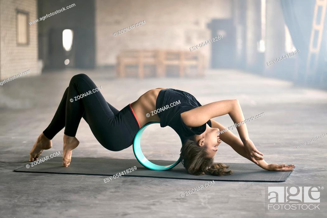 Imagen: Delightful girl with closed eyes in a black sportswear engaged in yoga in a loft style hall. She does a deflection while lies on the cyan yoga wheel and leans.
