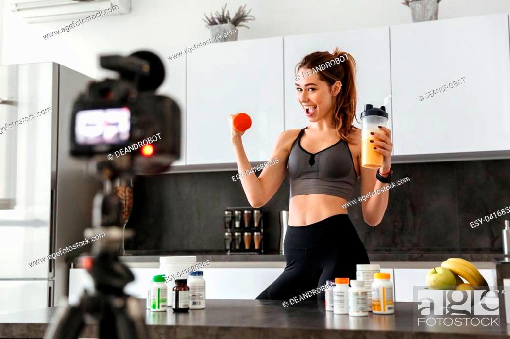 Stock Photo: Happy healthy young girl recording her video blog episode about healthy food additives while standing at the kitchen at home and holding dumbbell.