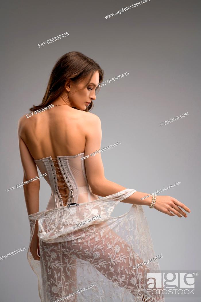 Stock Photo: Confident seductive slim female model in translucent corset keeping hands on waist and undress peignoir.