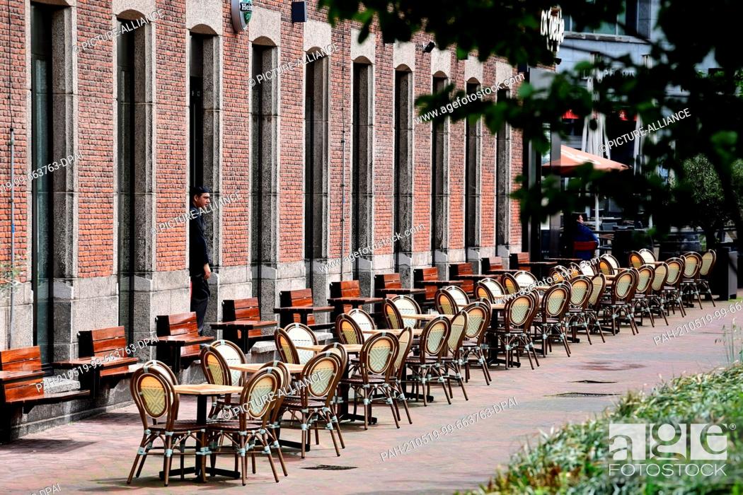 Stock Photo: 19 May 2021, Netherlands, Rotterdam: A waiter stands in sunny weather in one of the entrances in front of the empty tables and chairs of the restaurant.