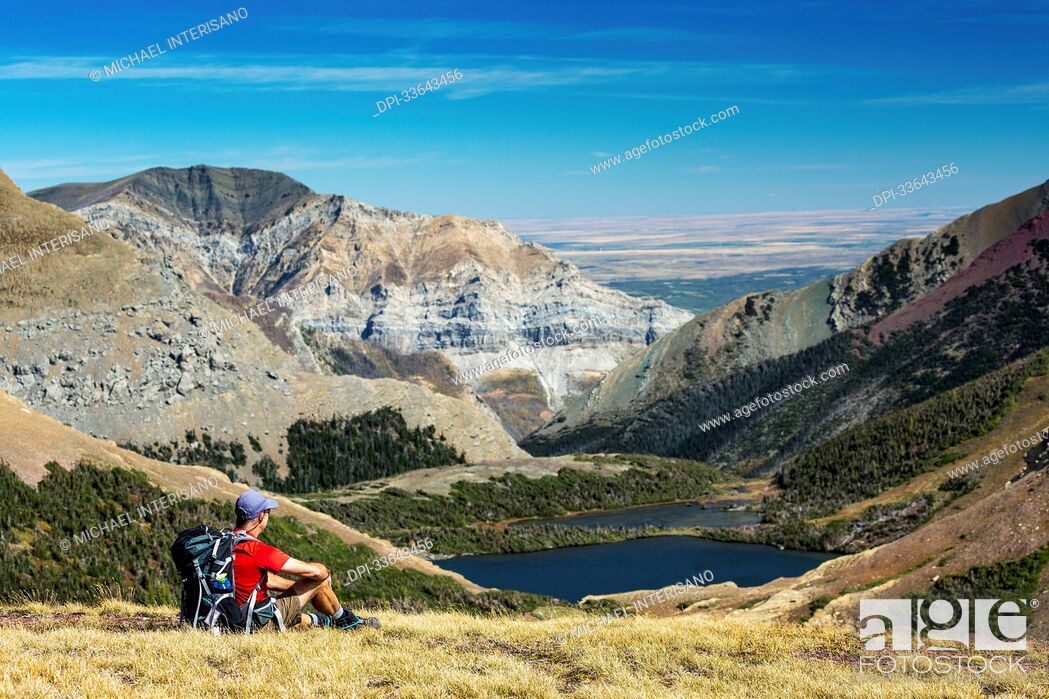 Stock Photo: Male hiker relaxing on grassy mountain ridge overlooking an alpine lake, mountain ranges, blue sky and clouds in the background.