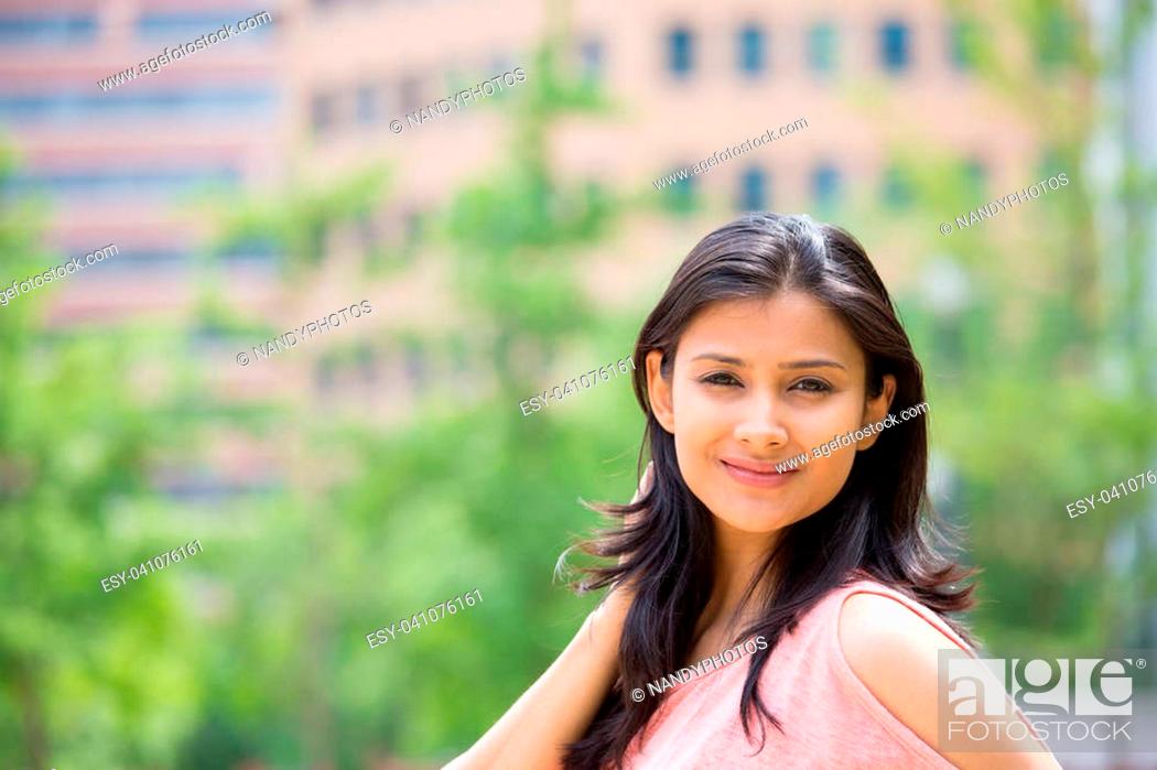 Stock Photo: Closeup portrait of confident smiling happy pretty young woman in pink dress, isolated background of blurred trees, buildings.
