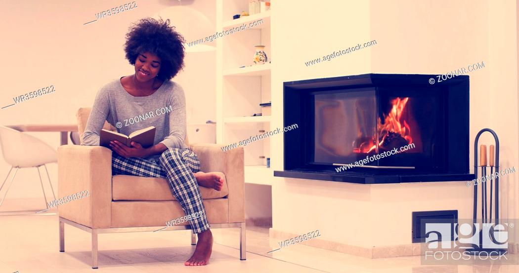 Stock Photo: Young beautiful african american woman sitting in front of fireplace at home on a cold autumn day and reading book.