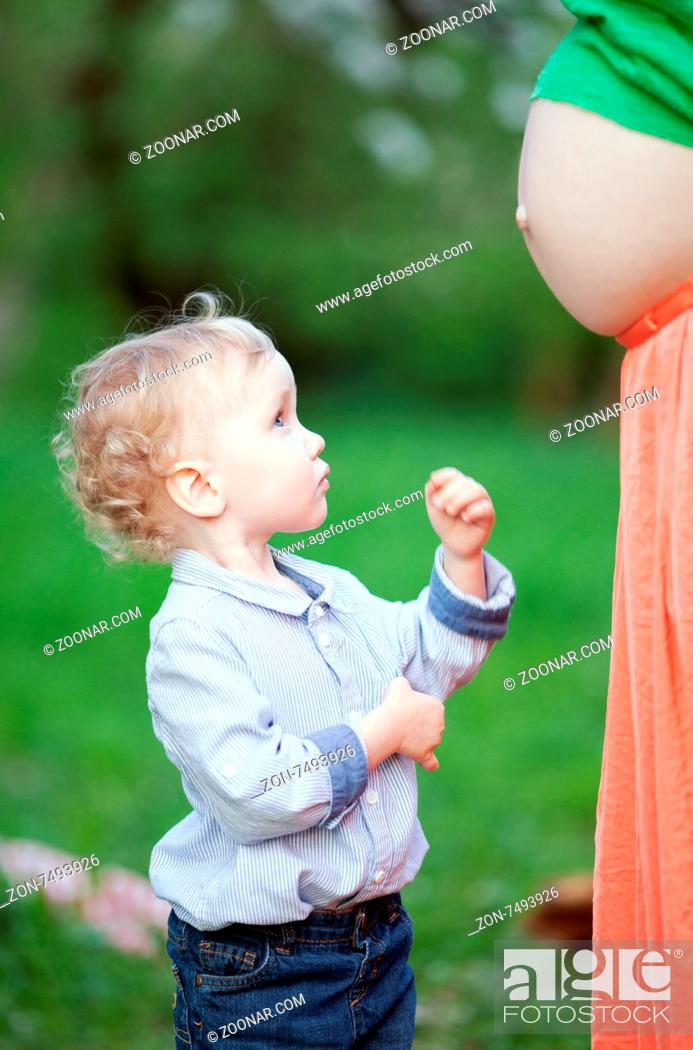 Stock Photo: Lovely little boy looking at pregnant mothers belly outdoor. He is expecting to have brother or sister.
