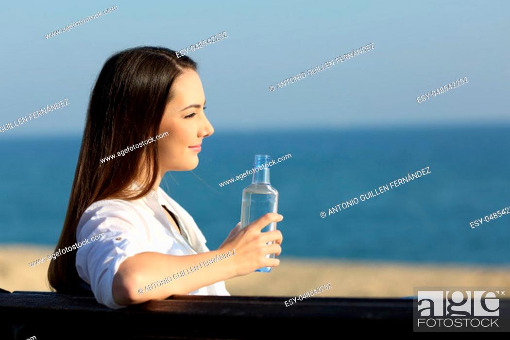 Stock Photo: Smiley woman holding a water bottle and looking at horizon on the beach.