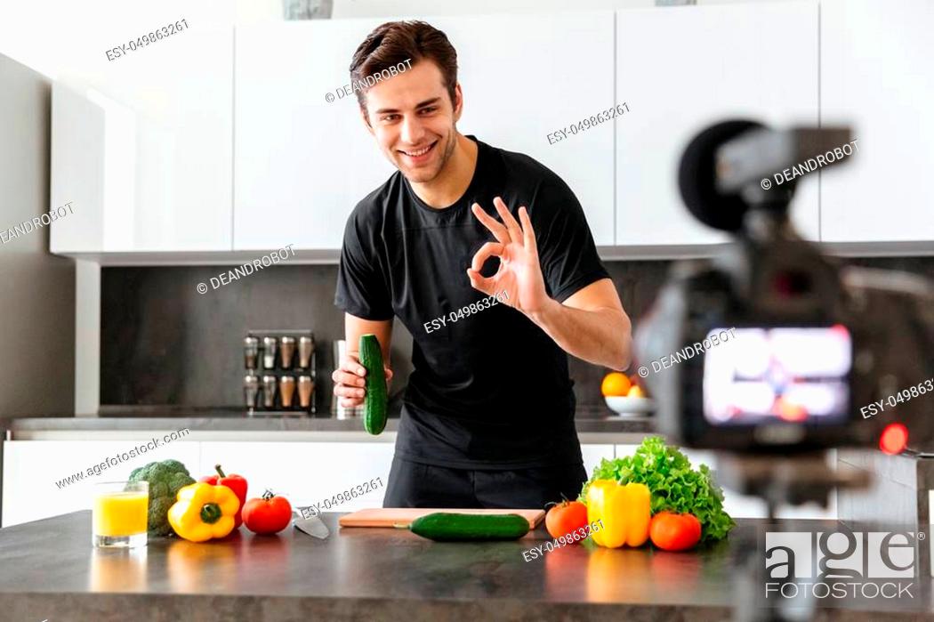Stock Photo: Smiling young man filming his video blog episode about healthy food cooking while standing and showing ok gesture at the kitchen.