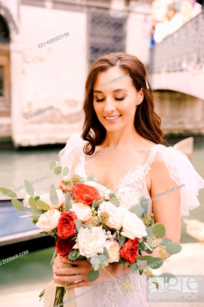 Stock Photo: A bride in a white dress, with a train, with a bouquet of white and red roses in her hands, stands on the pier near the moored gondola in a narrow Venetian.